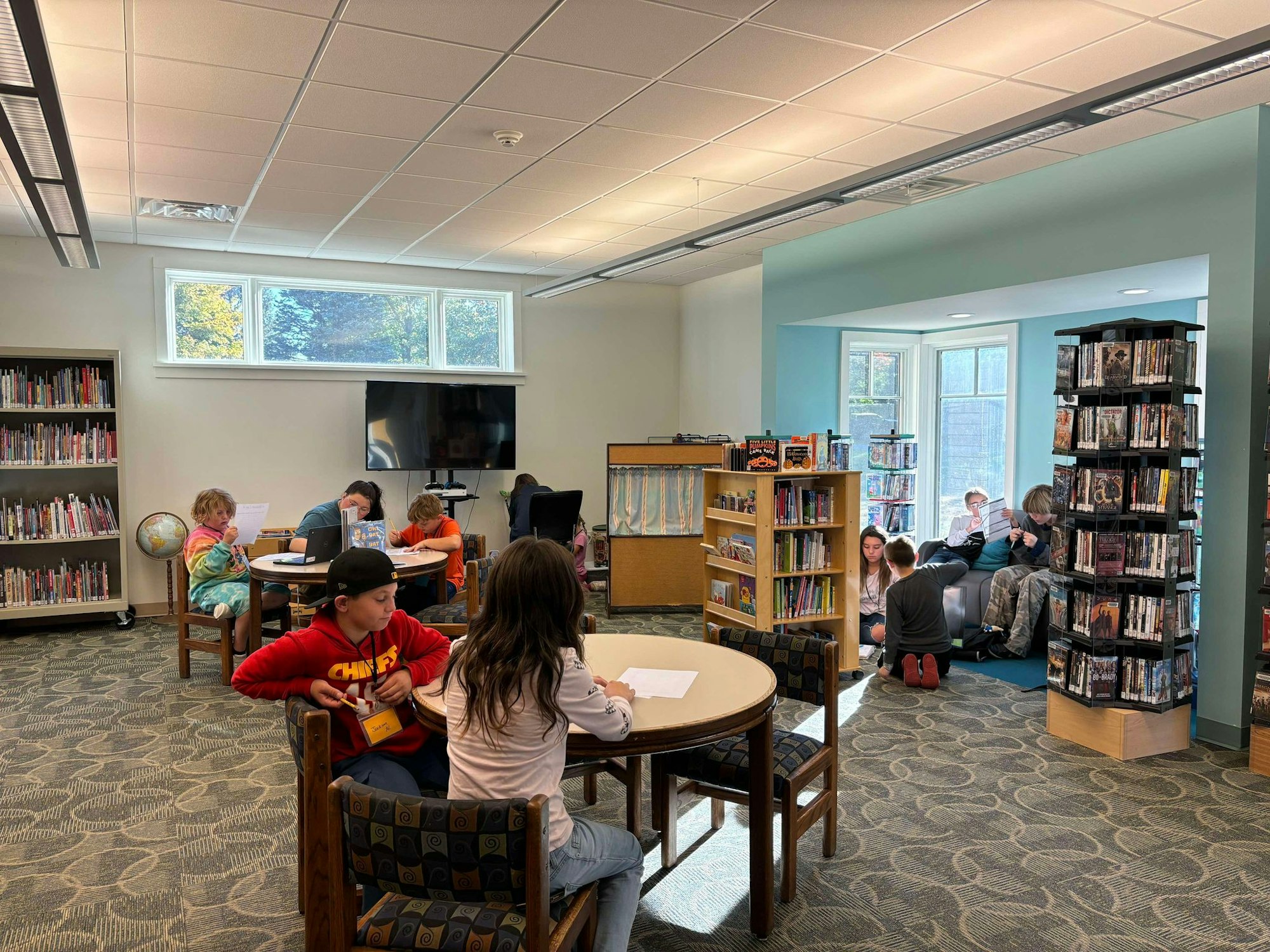 A bright library room with kids reading and using a computer, bookshelves, a TV, and a globe.