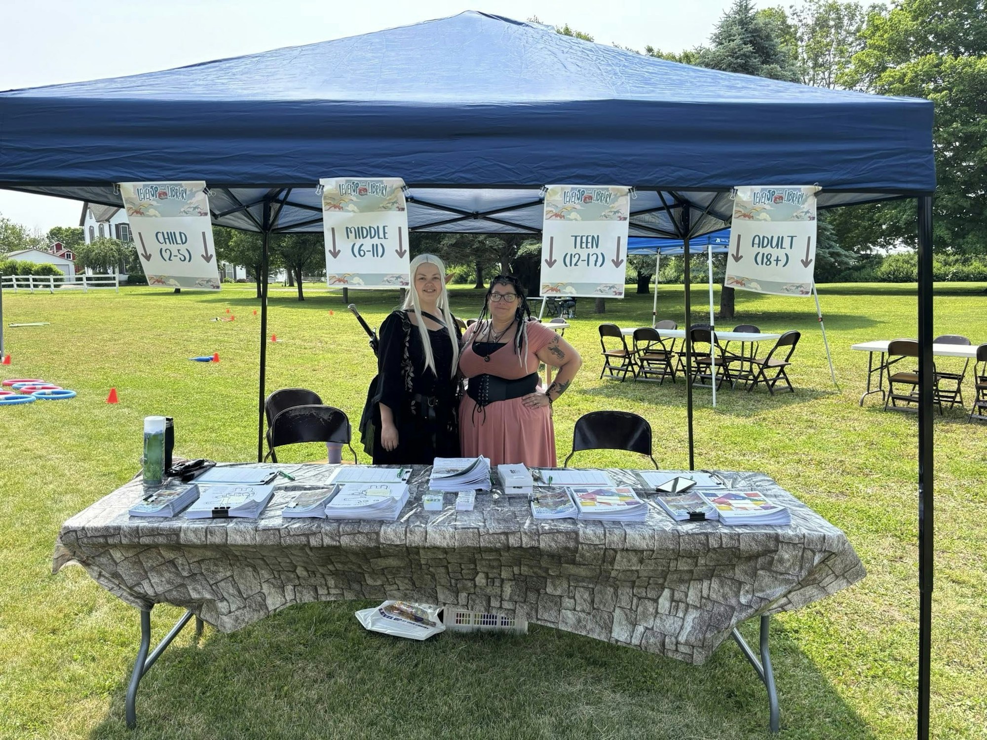 Two people at a booth under a canopy, with signs for age groups: Child, Middle, Teen, Adult. Outdoor setting with chairs and tables.
