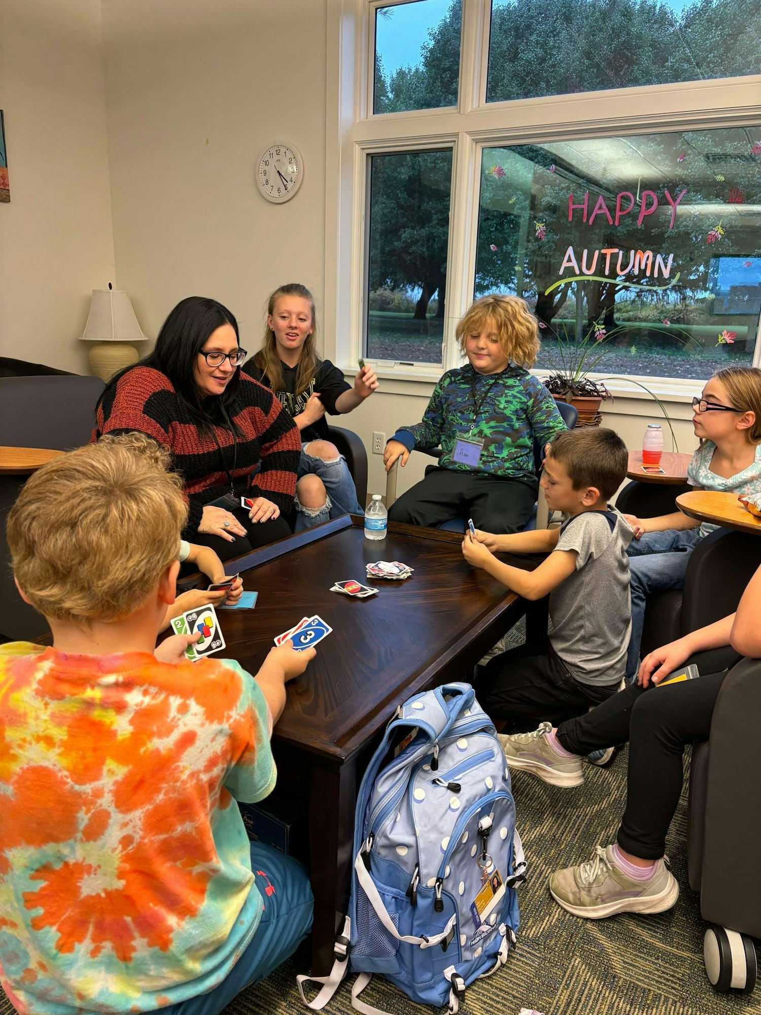 Group of kids and an adult playing a card game, "Happy Autumn" written on window, casual, fun atmosphere.