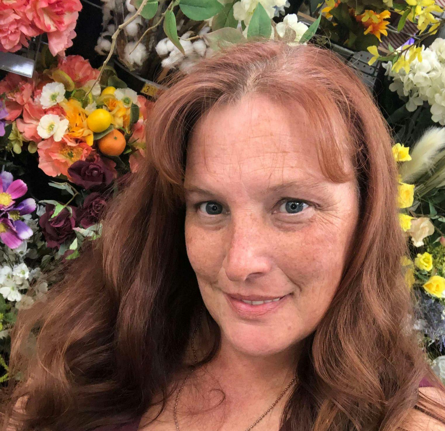 Selfie of a smiling woman with red hair, in front of a floral backdrop. She wears a purple top and a heart necklace.