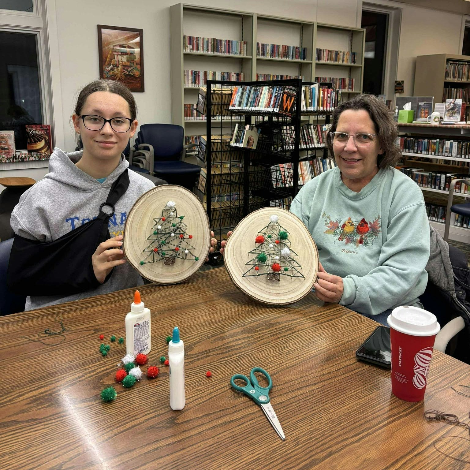 Two people display crafted Christmas tree decorations on wooden pieces, sitting at a table with glue, pom-poms, and scissors.