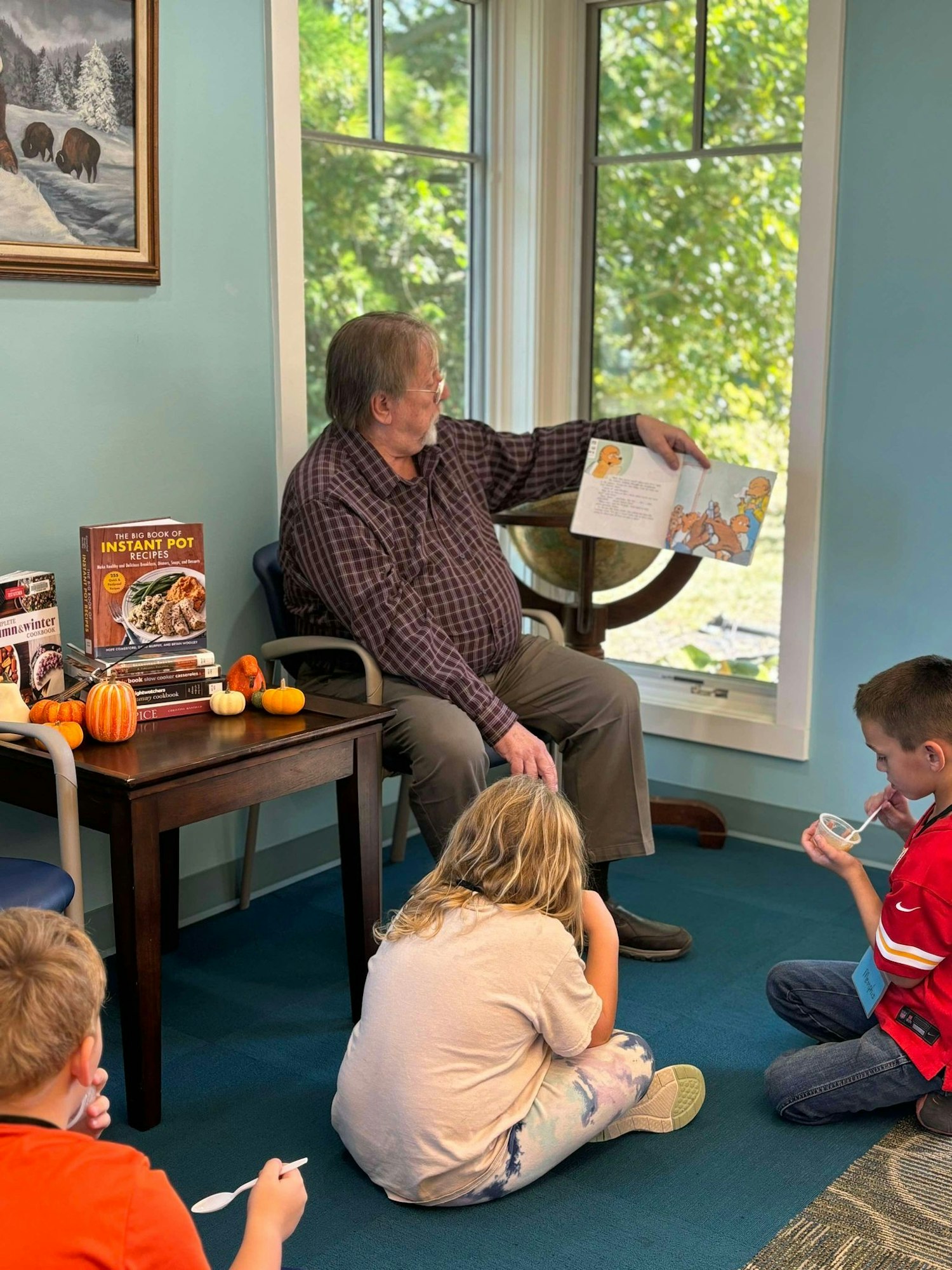 A person is reading to children indoors, with a book display and pumpkins on the table, a nature painting on the wall, and a scenic window view.