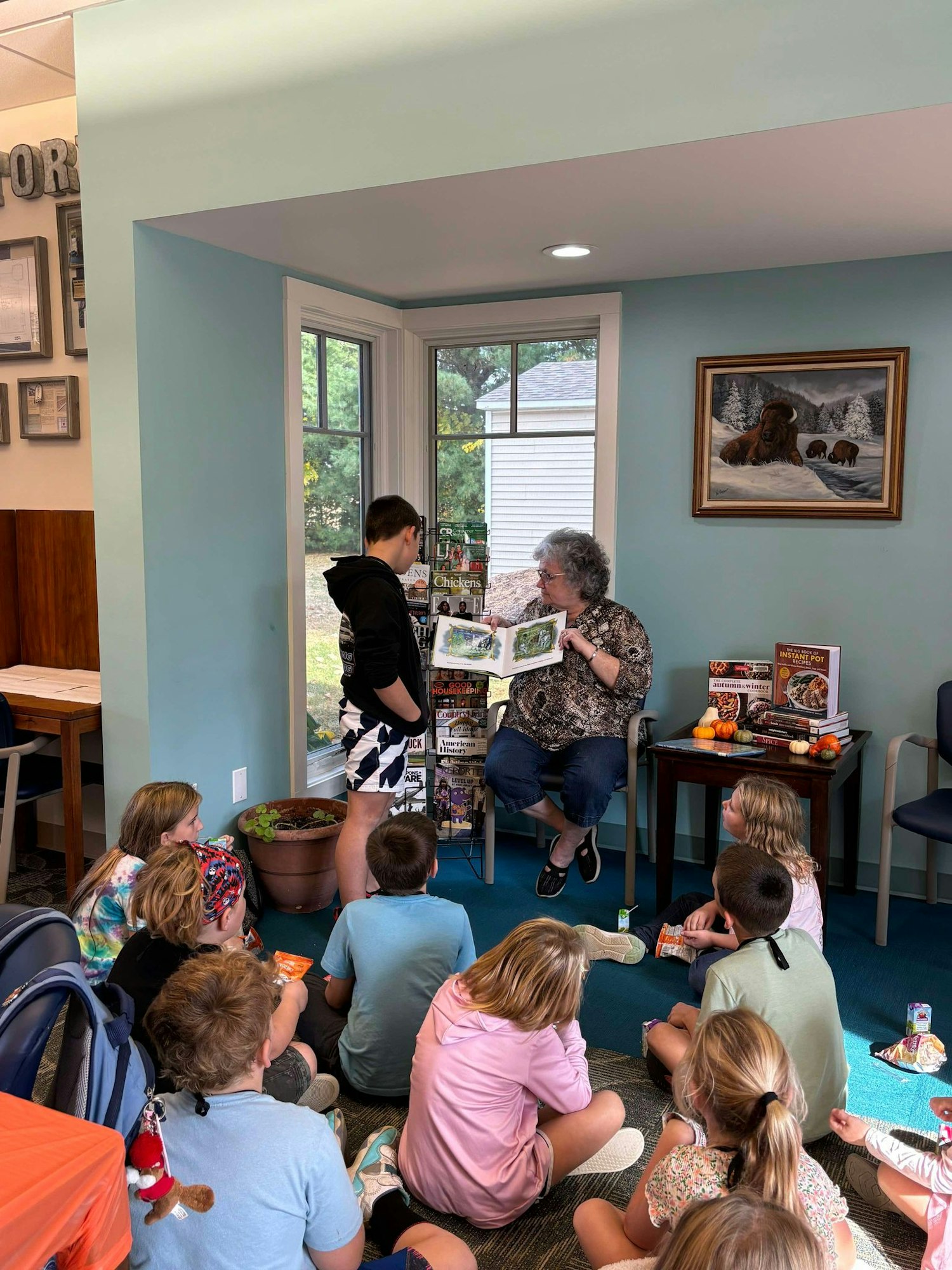 A group of children attentively listening to a story read by a seated woman, with another adult standing beside her, indoors.