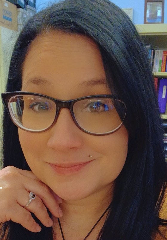 A smiling person with glasses, a nose piercing, and dark hair, possibly in a library.