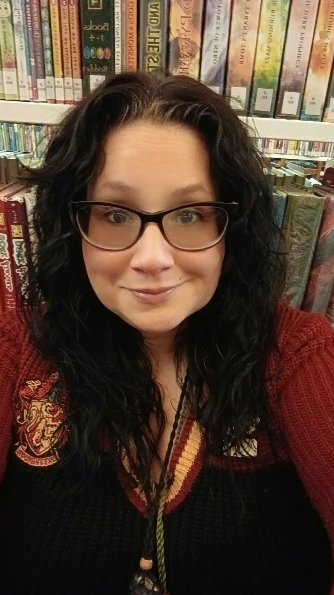 A woman with curly hair and glasses smiles at the camera, standing in front of a shelf filled with colorful books.