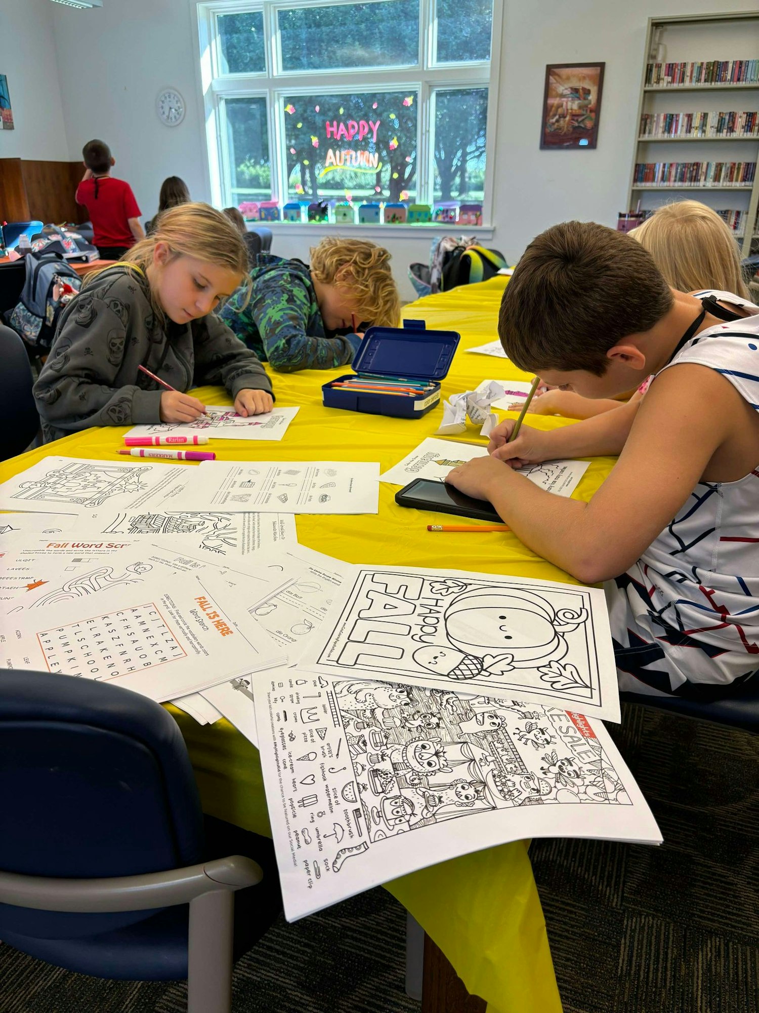 Children coloring and doing activities at a library with a "HAPPY AUTUMN" window decoration.