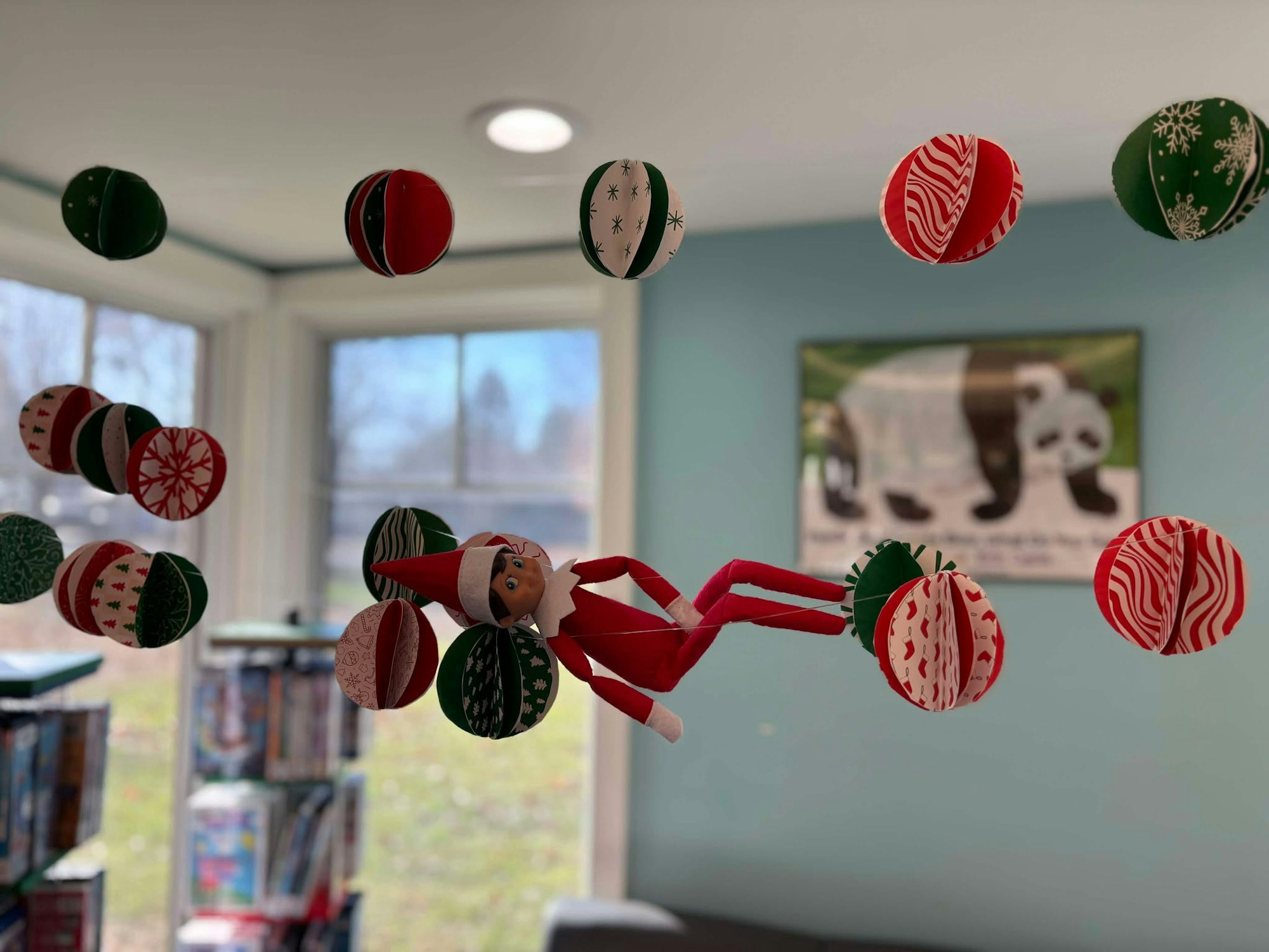 Elf on the Shelf among hanging Christmas-themed paper ornaments in a cozy room.