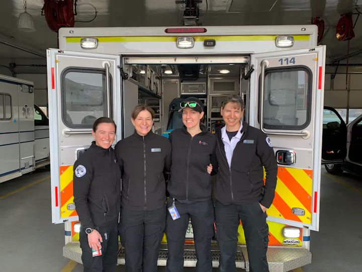 Four emergency responders pose together in front of an ambulance, showcasing teamwork in a professional setting.