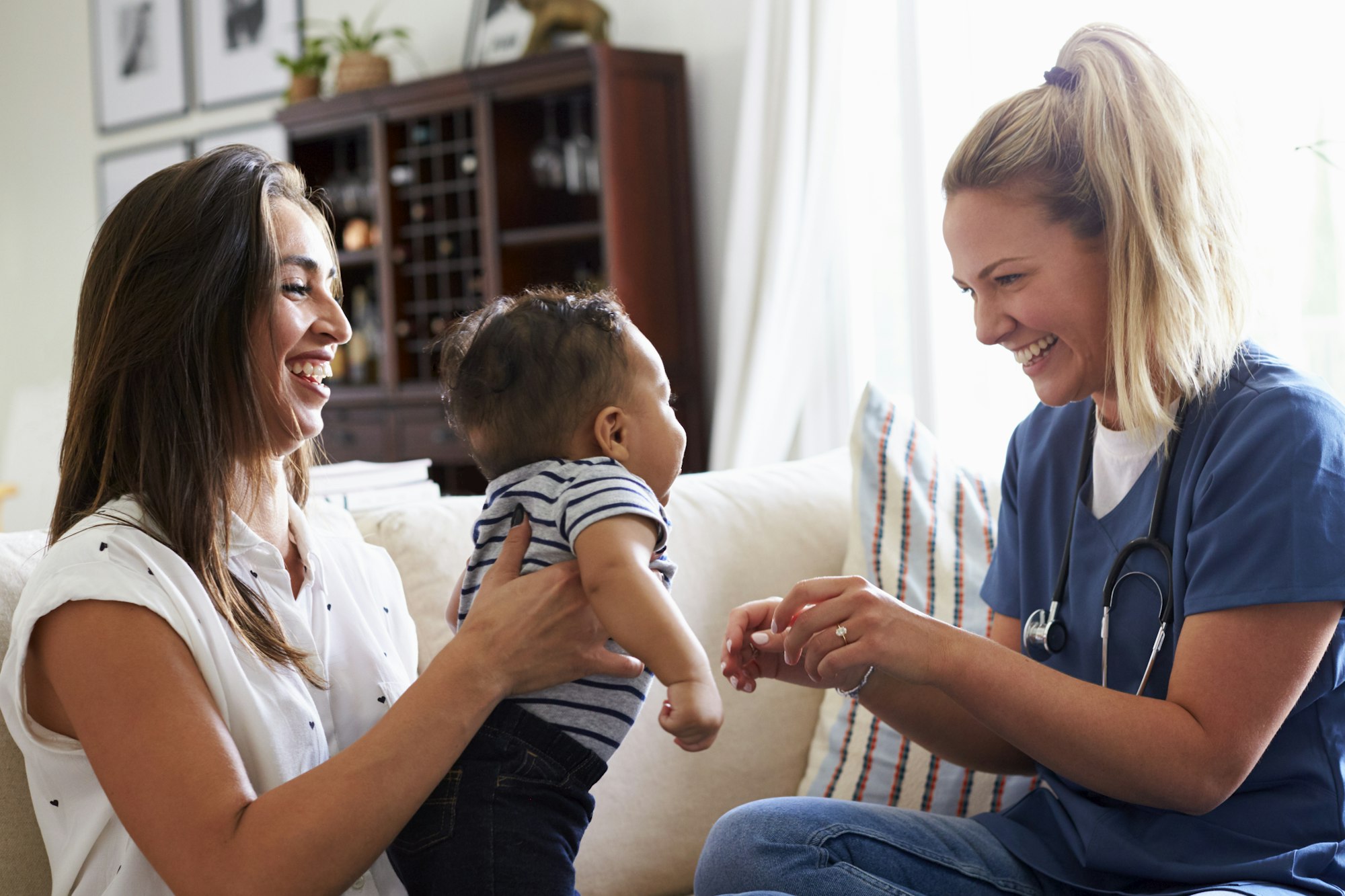 A woman holding a baby and a healthcare professional with a stethoscope are smiling and interacting.
