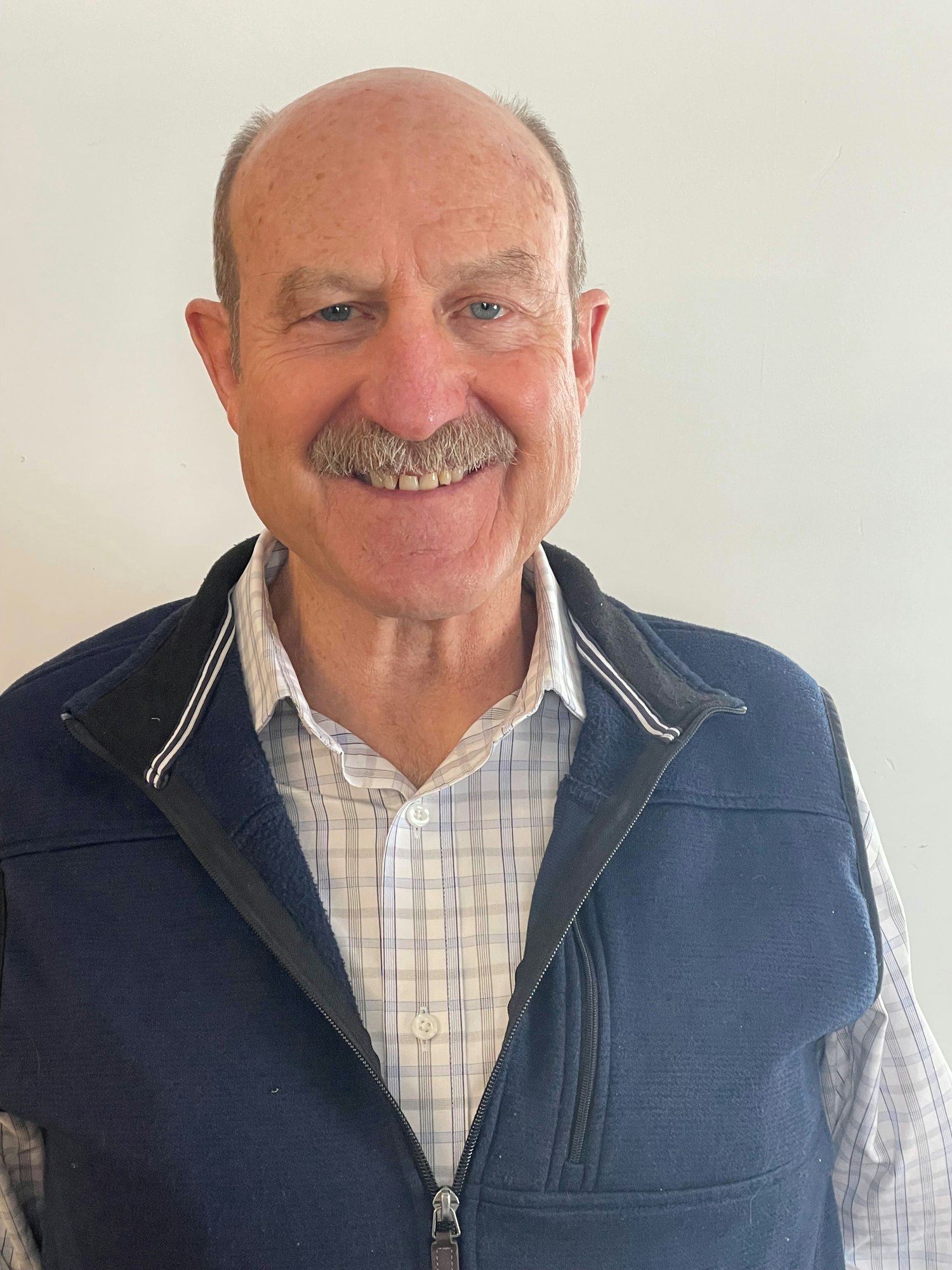 A smiling man in a checked shirt and dark vest against a plain background.