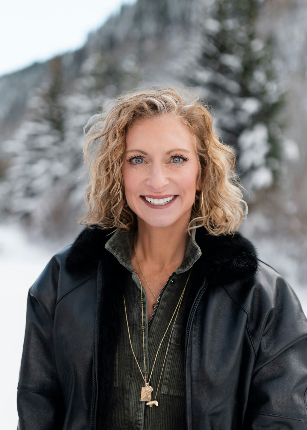 A person wearing a black jacket and necklace stands outdoors with a snowy forest background.