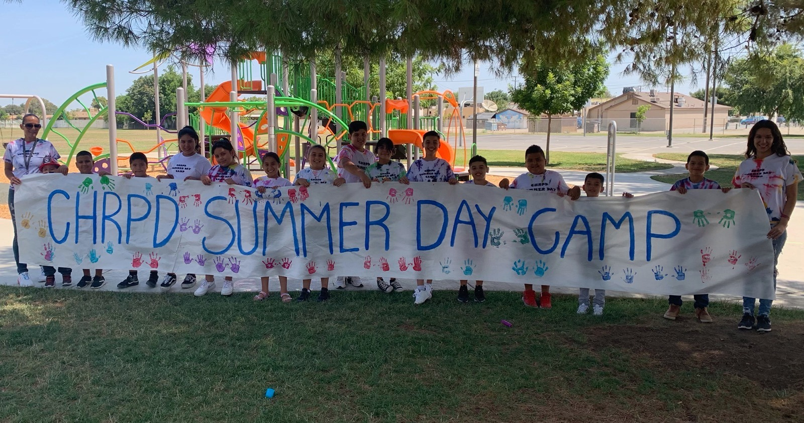 Group of children and adults holding a "CHRPD SUMMER DAY CAMP" banner at a park with a playground in the background.