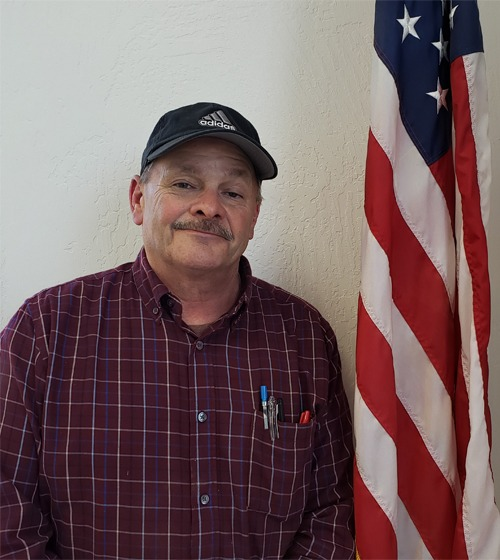 A man in a cap and plaid shirt smiling, with an American flag in the background.