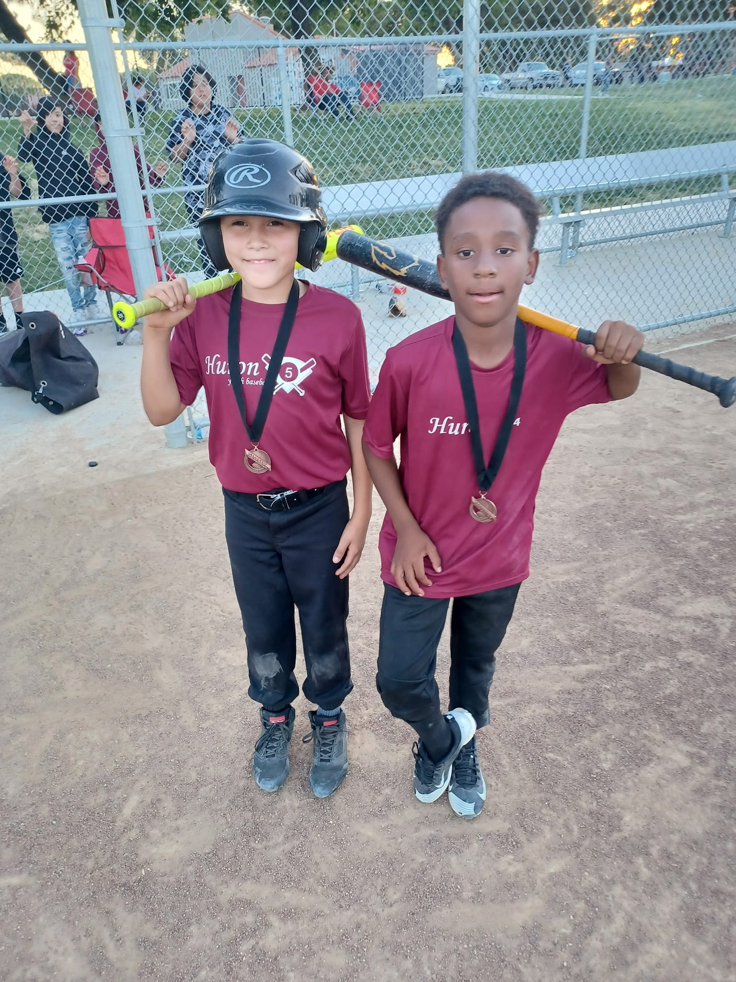 Two kids in baseball uniforms with medals and bats, standing on a dusty field, behind a fence with spectators.