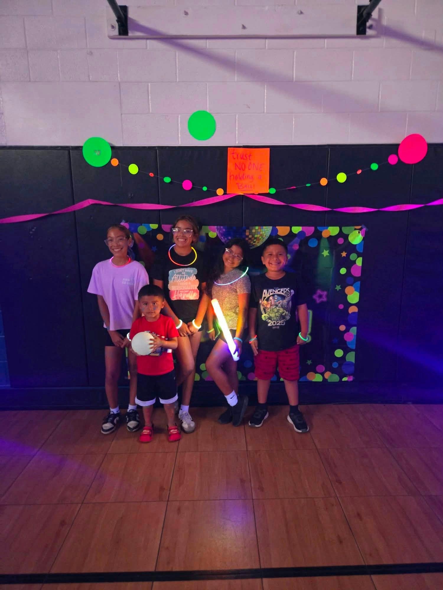 A group of five kids in a gym poses under colorful lights and decorations, holding glowing accessories and having fun.