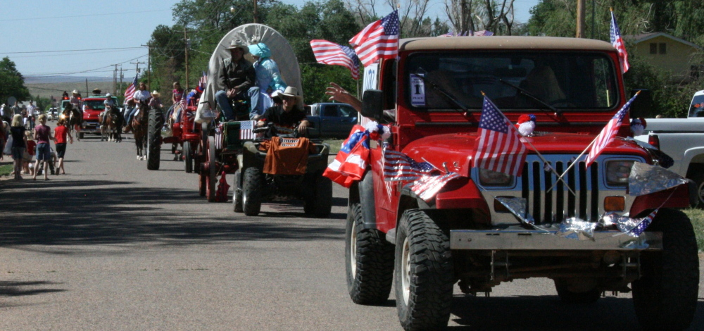 A parade with decorated vehicles and spectators, featuring American flags and a covered wagon.