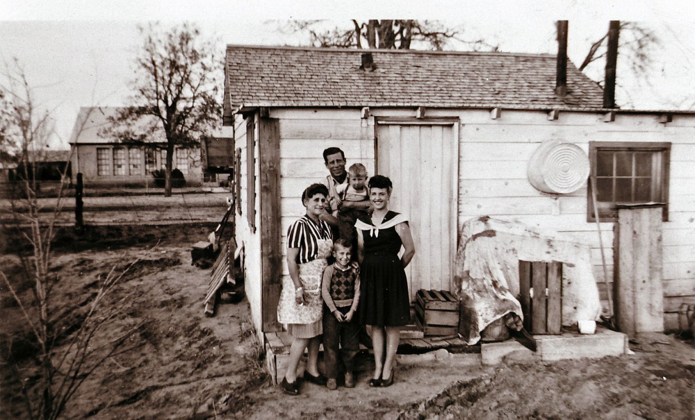 A vintage photo of five people posing in front of a rustic small house with sparse surroundings.
