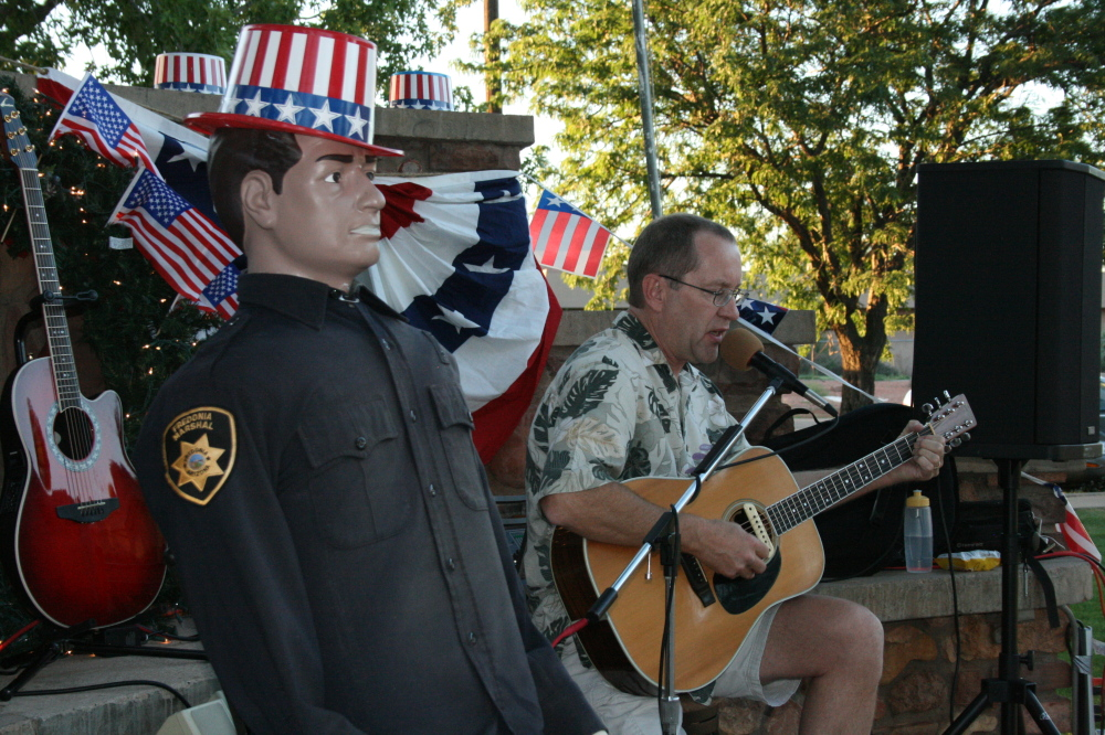 A man plays guitar beside a life-sized figurine in a sheriff's uniform. American flags and patriotic hats adorn the background.