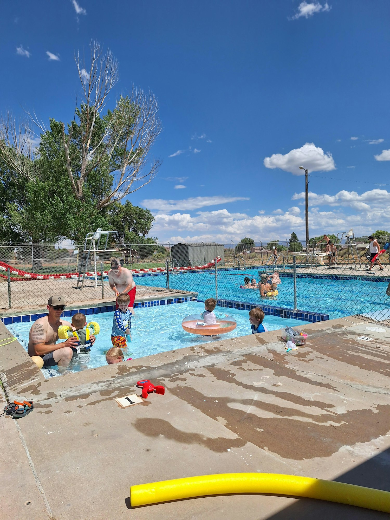 A crowded outdoor kiddie pool with people swimming and relaxing, under a blue sky with a few clouds.