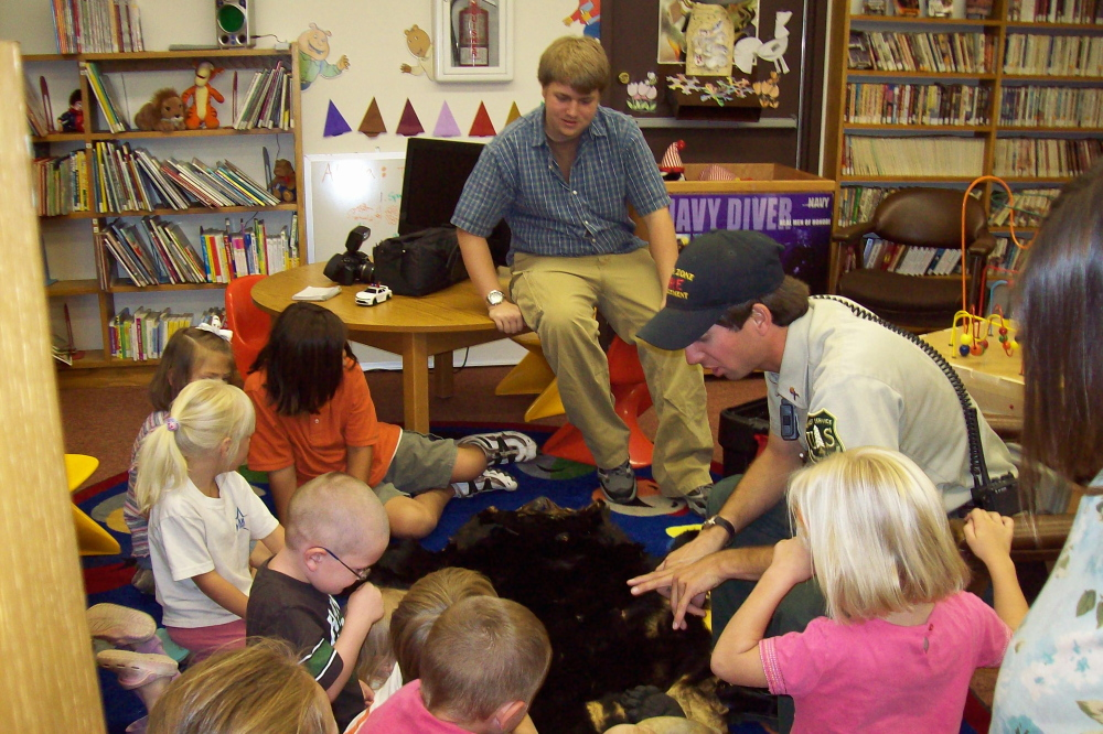Children interact with a dog during a show-and-tell session, with adults supervising in a library setting.