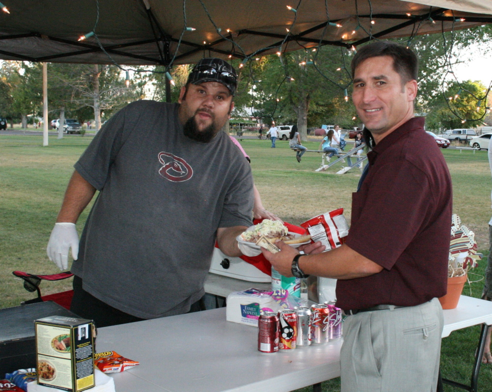 Two men at an outdoor event, one serving food and the other holding a plate and snacks, with people in the background.