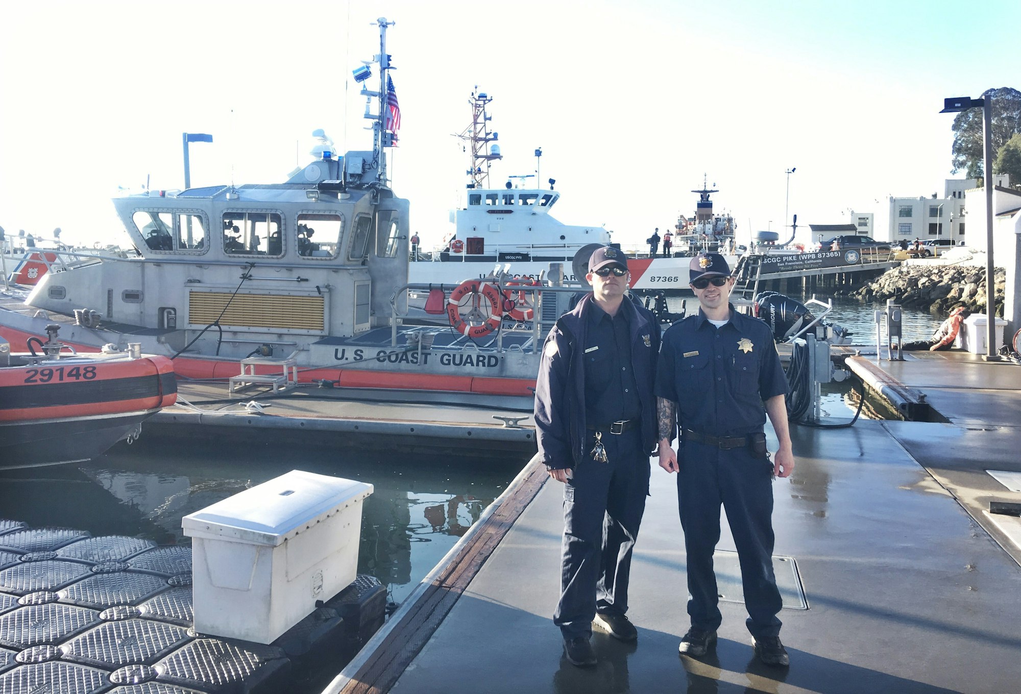 Two Coast Guard personnel stand near their boats at a harbor, showcasing their uniform and equipment.