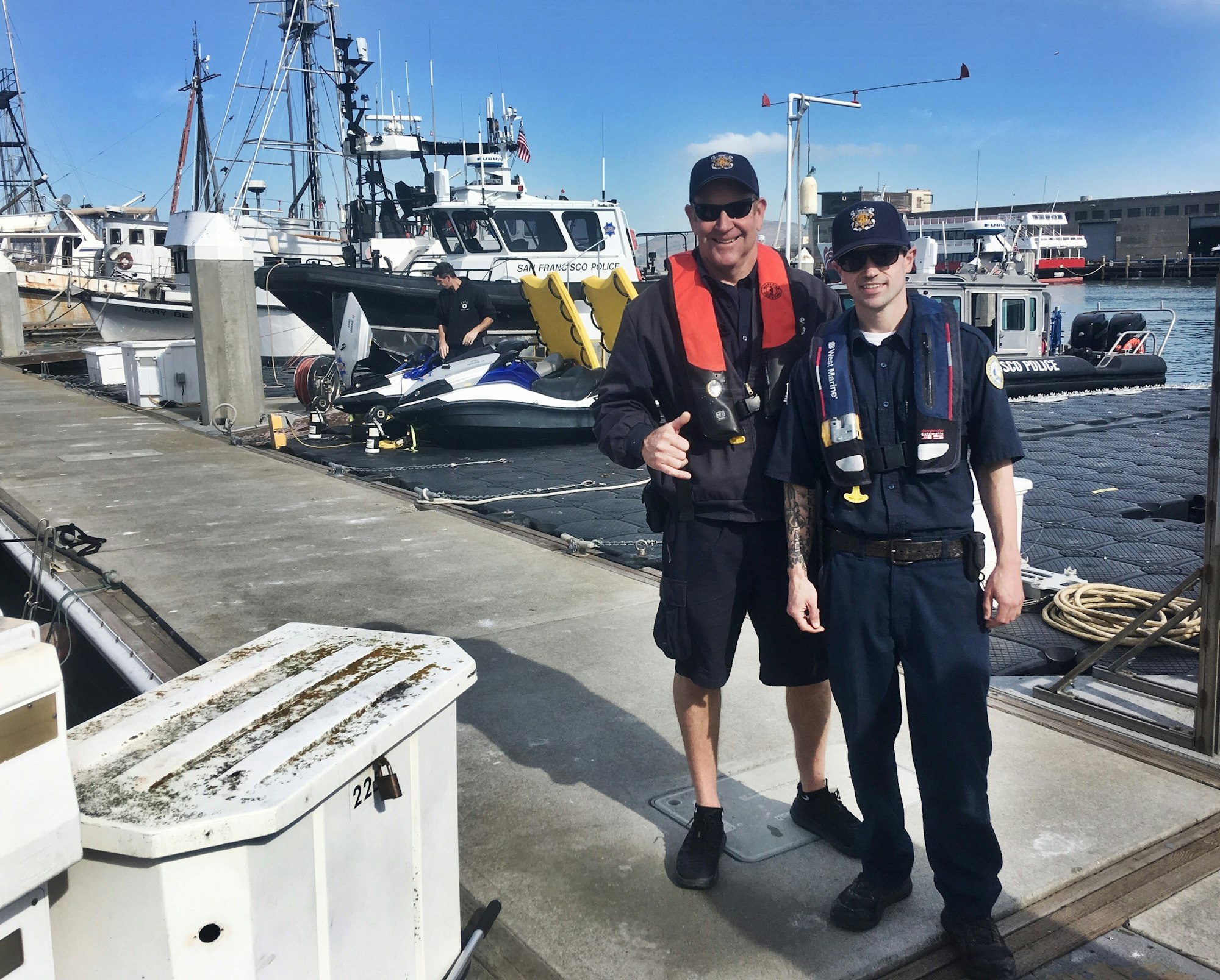 Two uniformed officers pose at a marina with police boats and a jet ski in the background on a sunny day.