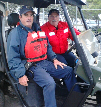 Two men in uniforms are seated in an all-terrain vehicle, wearing safety vests, ready for work.