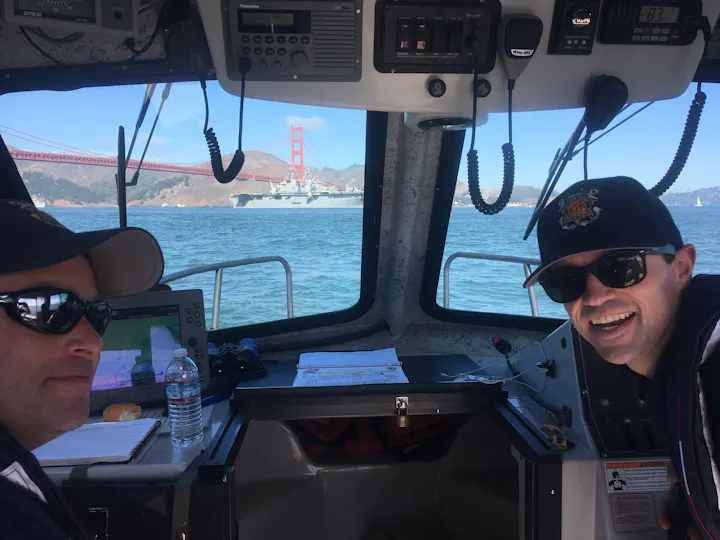 Two people are on a boat with views of the Golden Gate Bridge and a ship in the background, enjoying a clear day on the water.