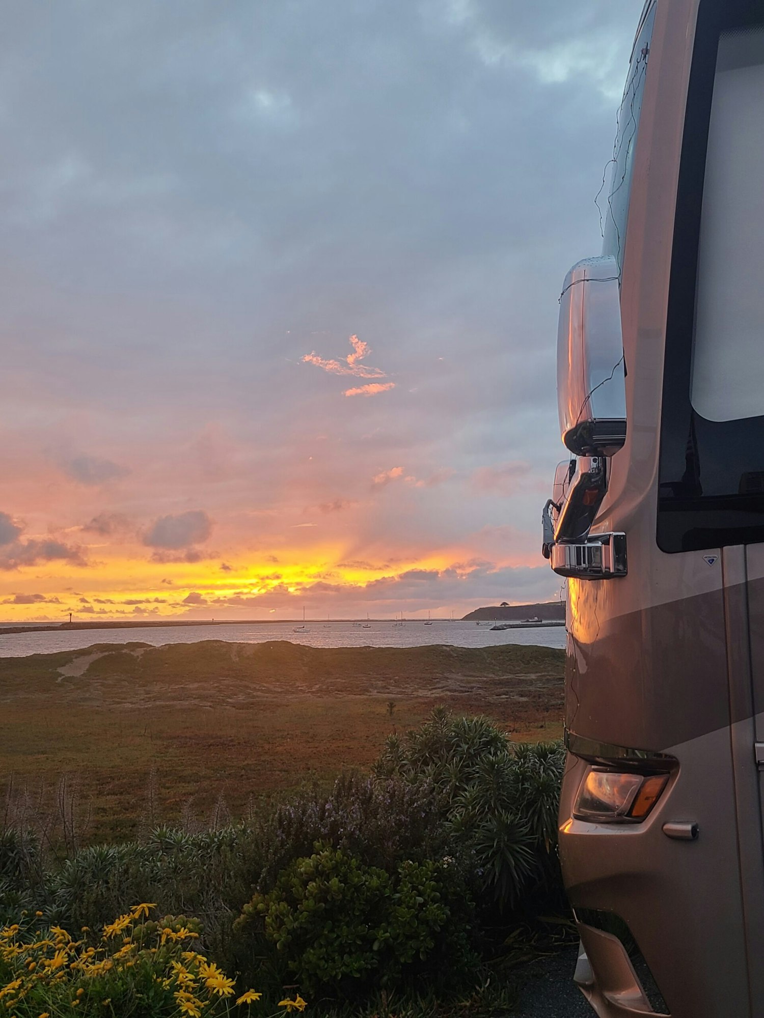 A camper van parked by the coast, with a scenic sunset over the ocean, boats, and flowers in the foreground.