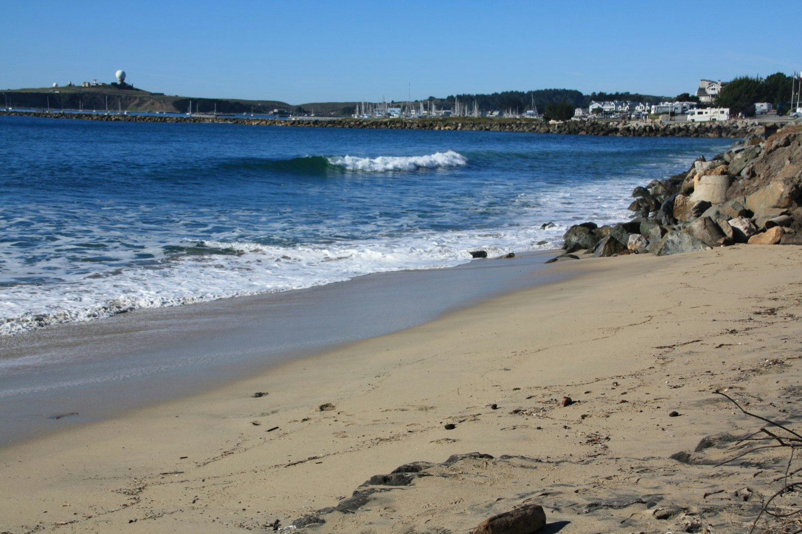 A sandy beach with gentle waves, rocks, and distant hills under a clear blue sky.