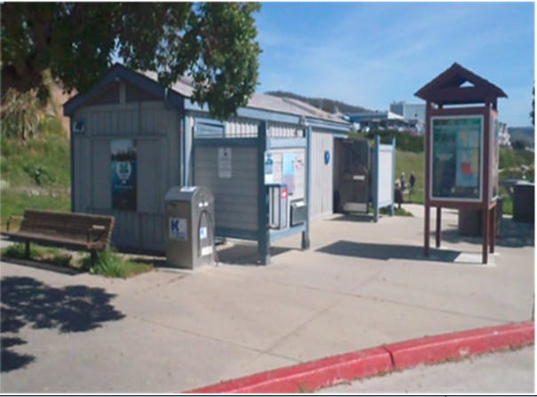 The image shows a public bus stop area with a shelter, bench, informational signs, and a recycling bin.