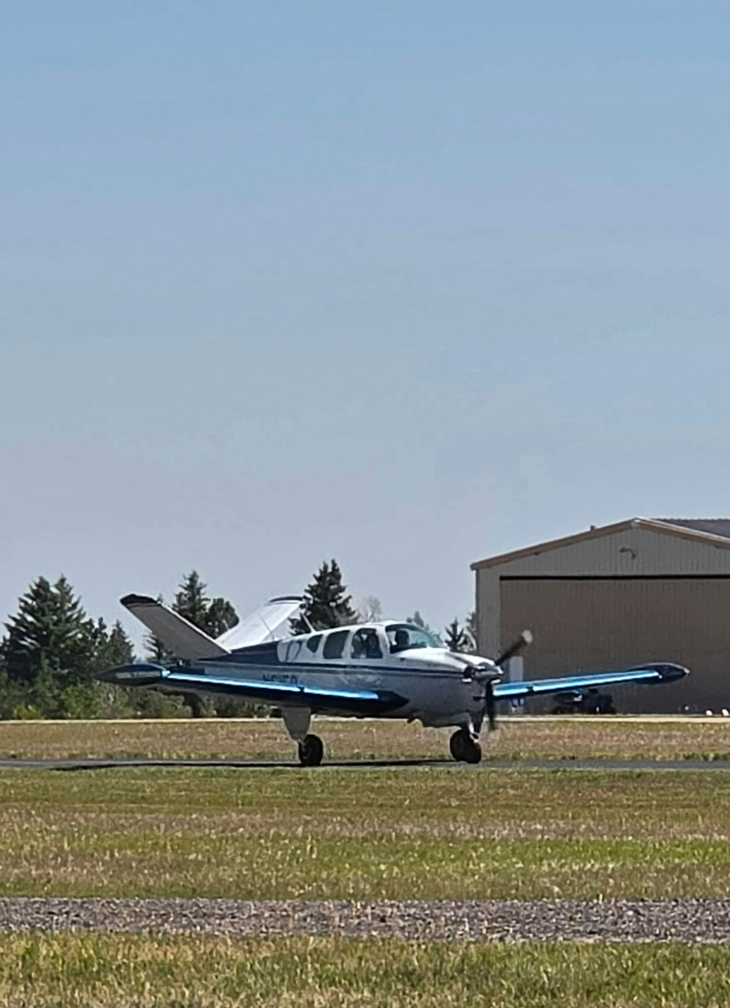 A small airplane with blue accents is parked on a runway near a hangar, surrounded by grass and trees under a clear sky.