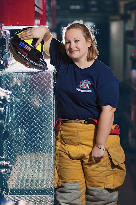 A smiling firefighter standing next to a fire truck, holding a helmet.