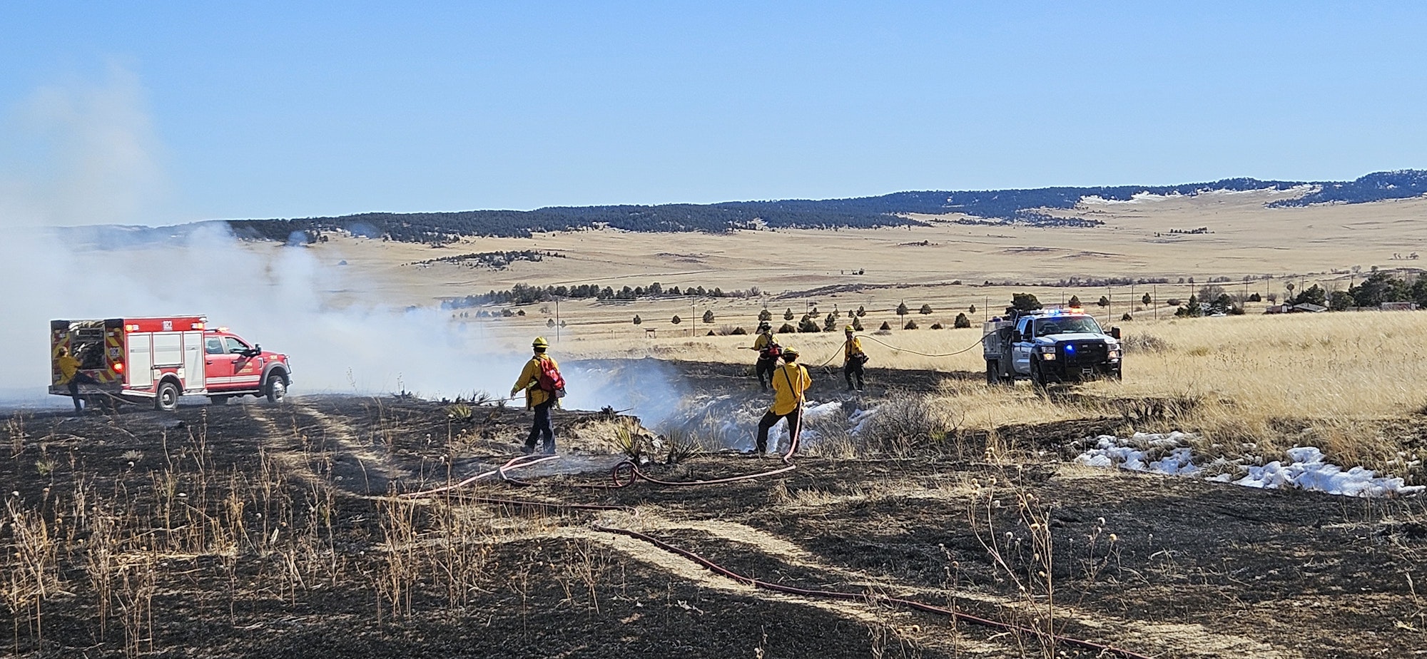 Firefighters battling a grass fire with trucks and hoses, amidst a smoke-filled landscape.