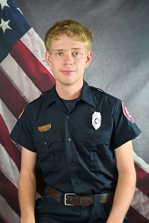 A young man in a police uniform poses in front of a backdrop featuring an American flag.