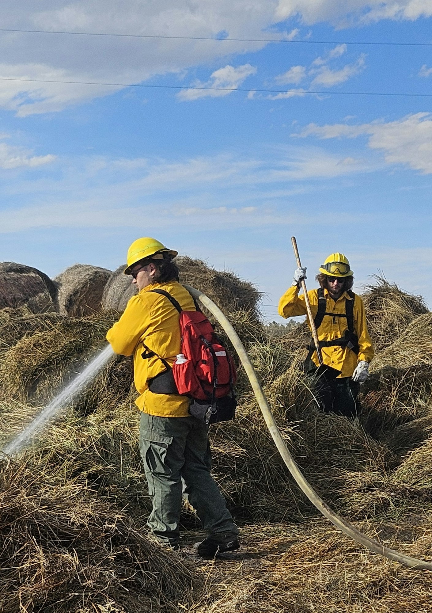 Two firefighters extinguishing a hay stack, clear skies in the background.