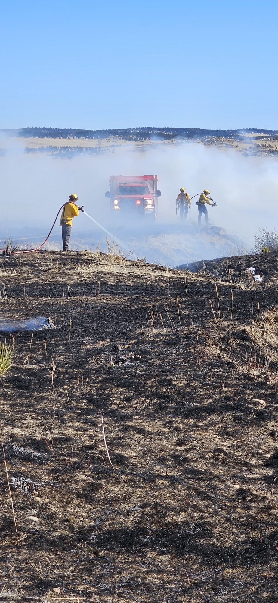 Firefighters extinguishing a field fire near a firetruck amidst smoke.