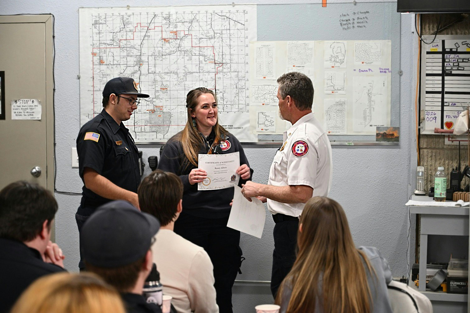 A ceremony where a woman receives a certificate of achievement, attended by colleagues in a training or meeting space.