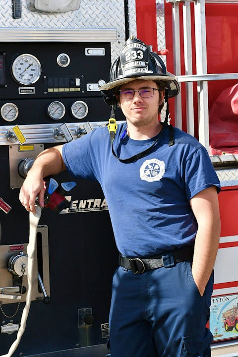 A firefighter in uniform stands by a fire truck, leaning casually against the controls.
