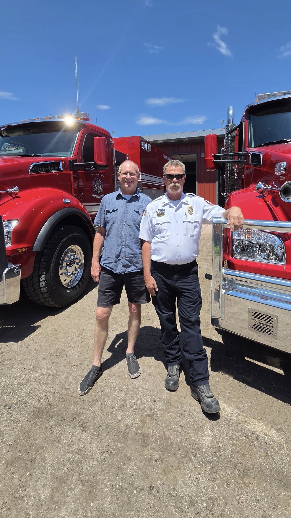 Two men pose with two large red trucks in the background on a sunny day. One man is in a uniform.