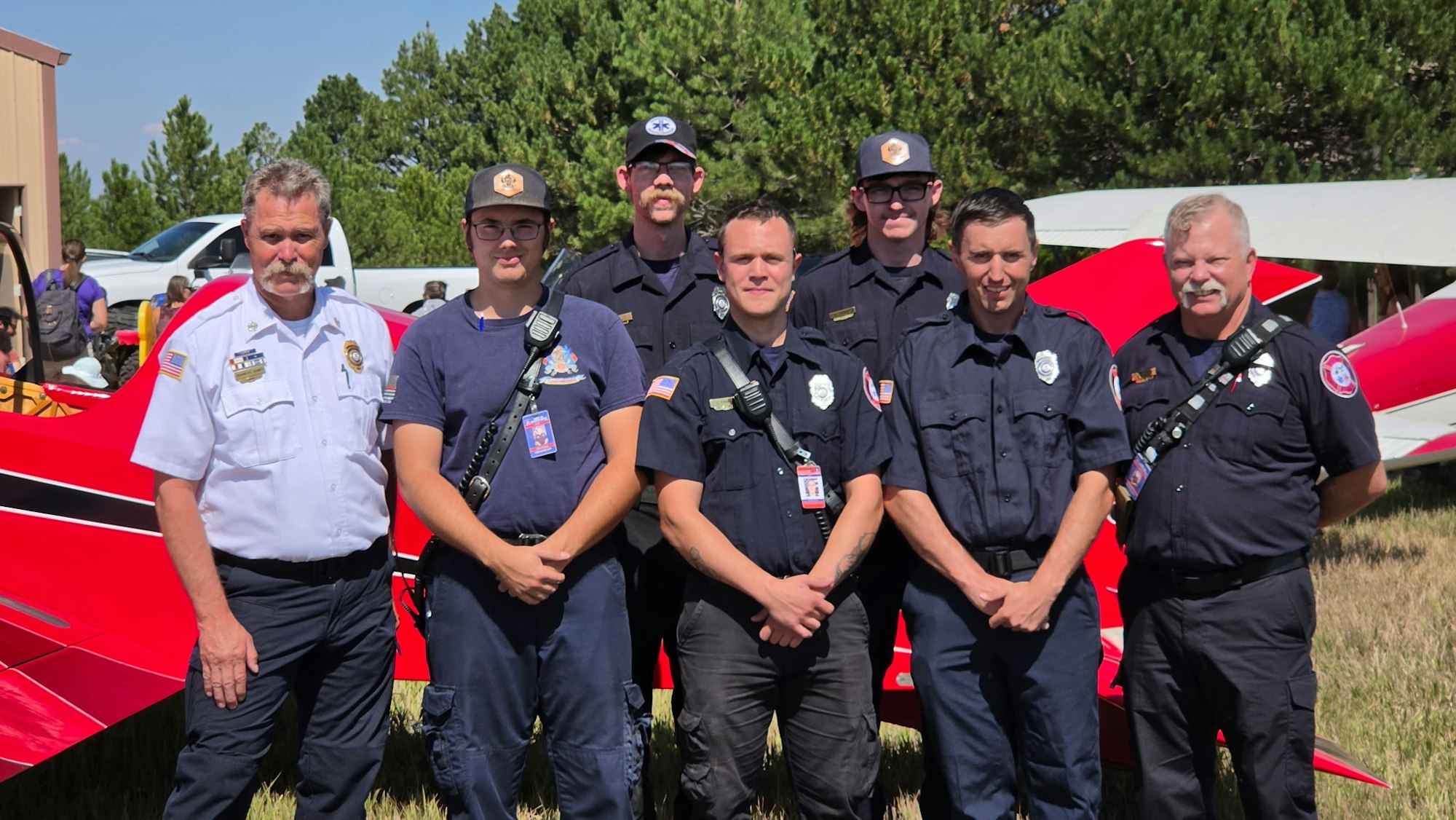 A group of six uniformed personnel poses in front of a red aircraft, likely at an event or training.