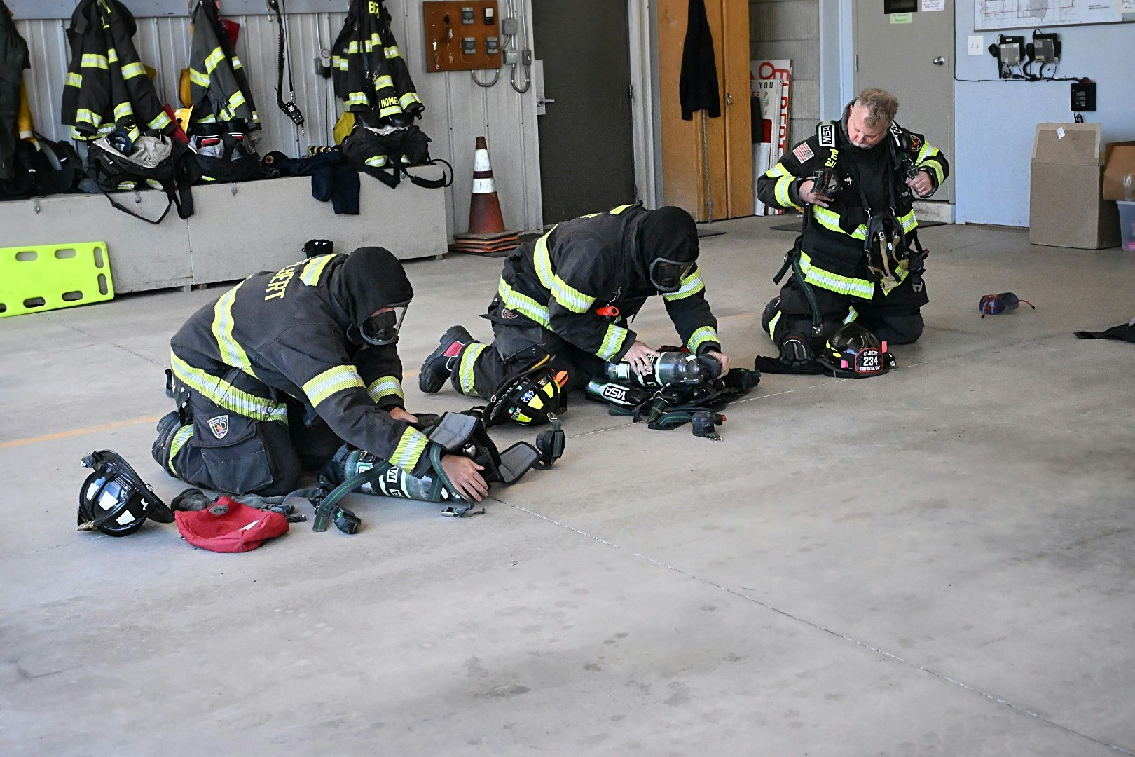 Firefighters are kneeling on the floor, checking equipment and wearing protective gear in a fire station.