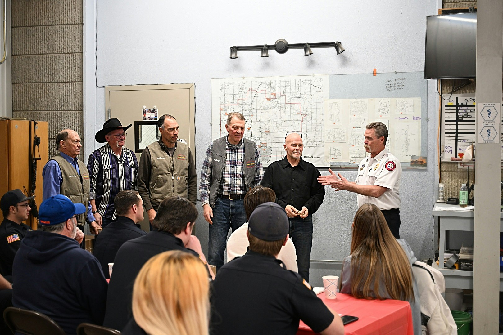 A gathering with several individuals at the front, likely for an event or presentation, with a map on the wall behind them.