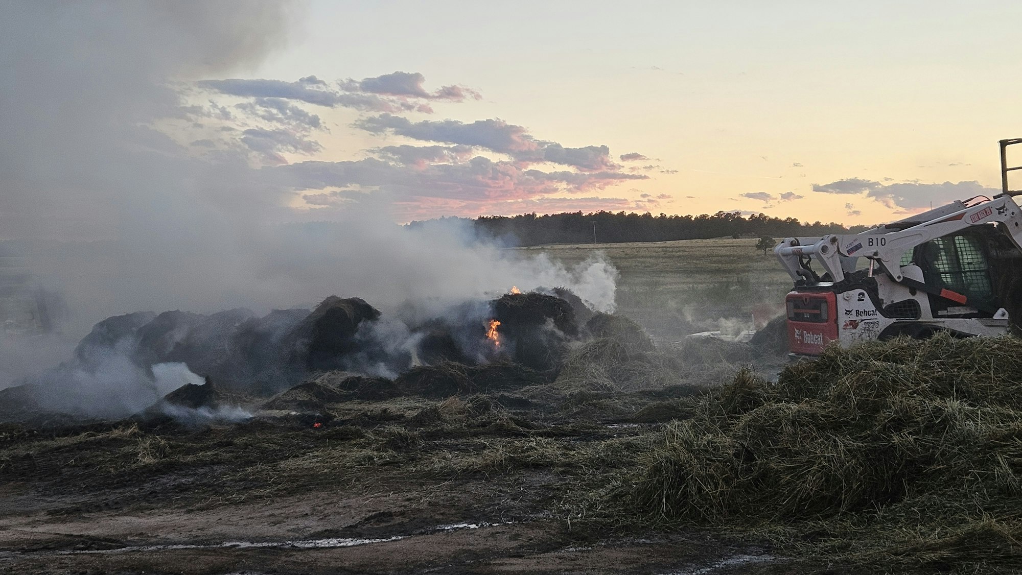 A smoldering pile of hay with a skid-steer loader nearby at dusk. Smoke rises against a cloudy sky.