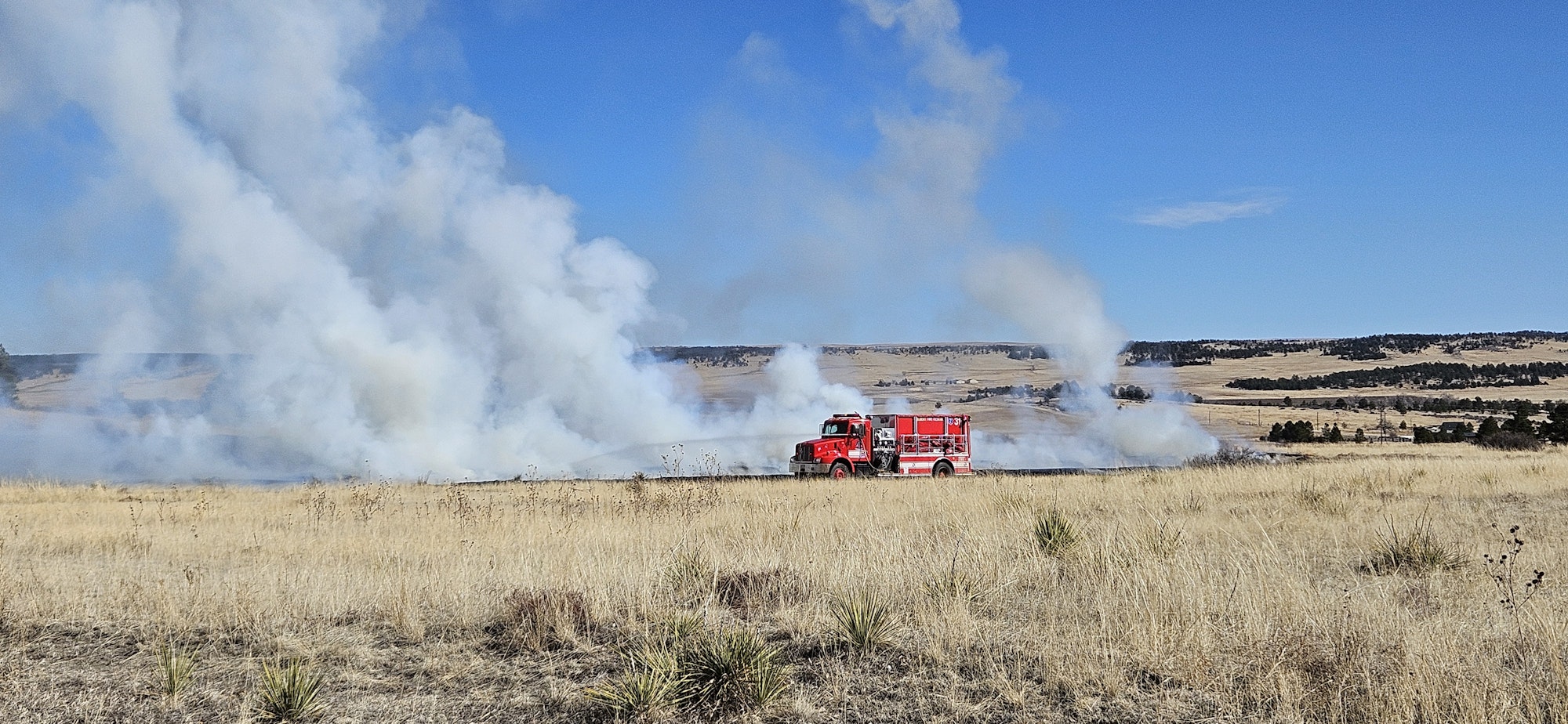 A fire truck is in a field with smoke, suggesting a wildfire event or controlled burn.