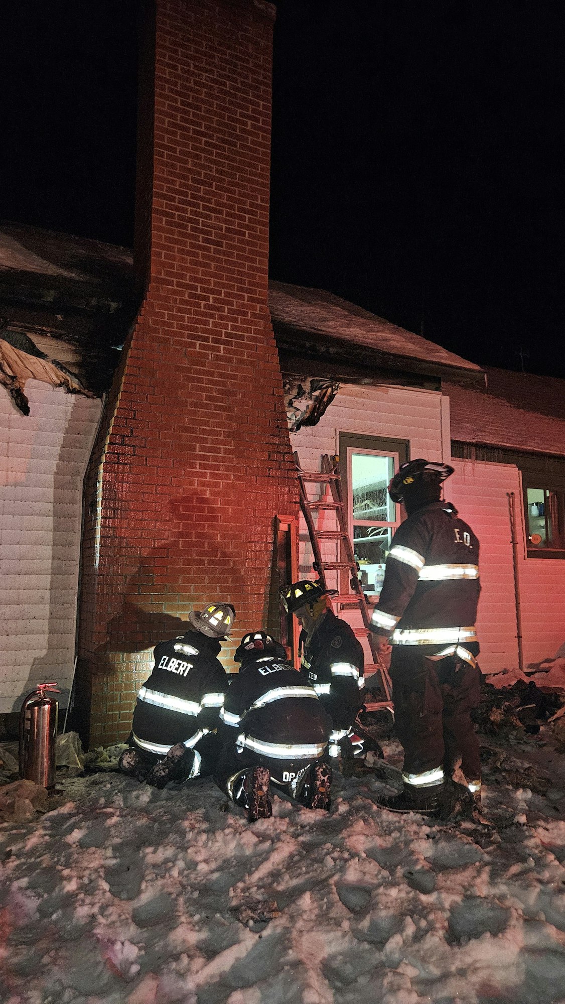 Firefighters in gear inspecting a building at night, with a ladder against the house and snow on the ground.