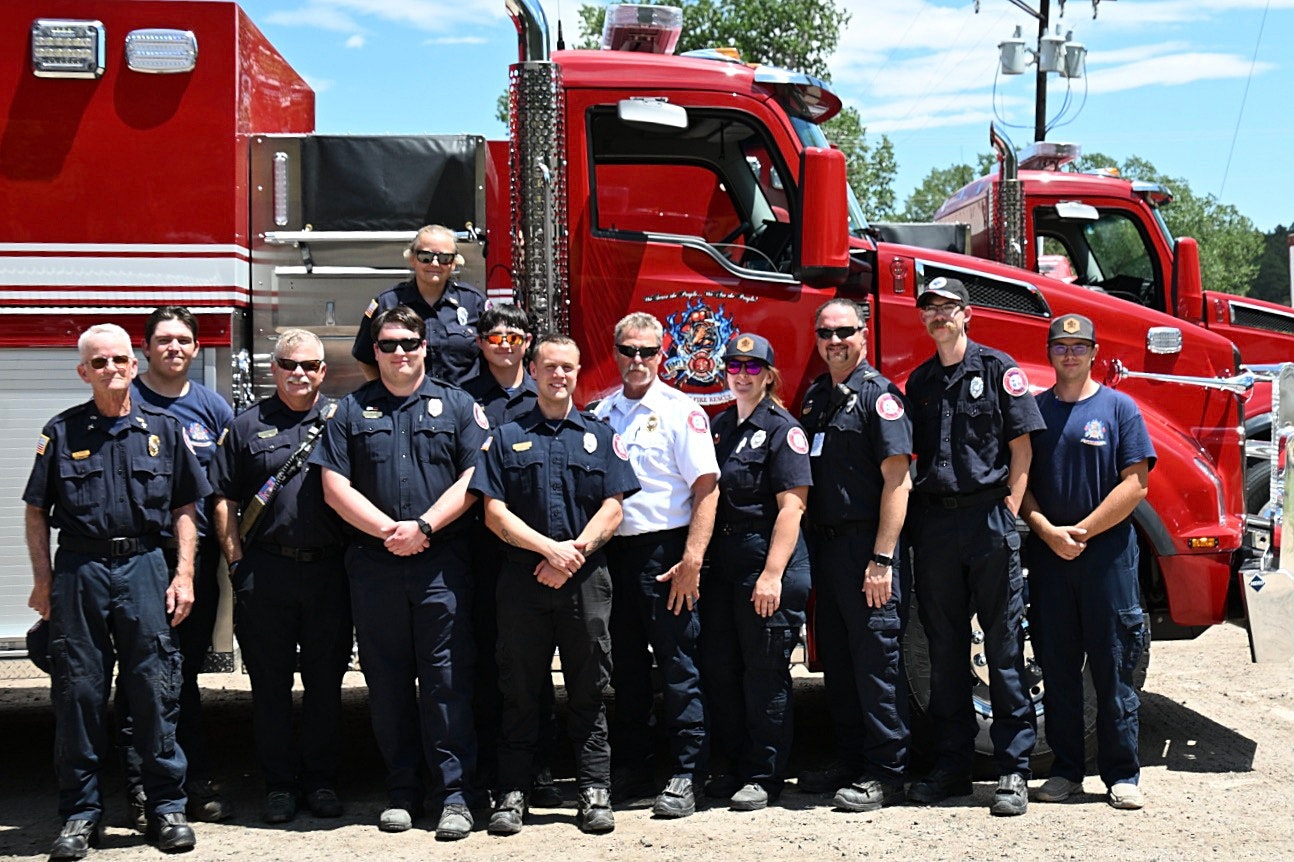 A group of firefighters posing together in front of a red firetruck on a sunny day.