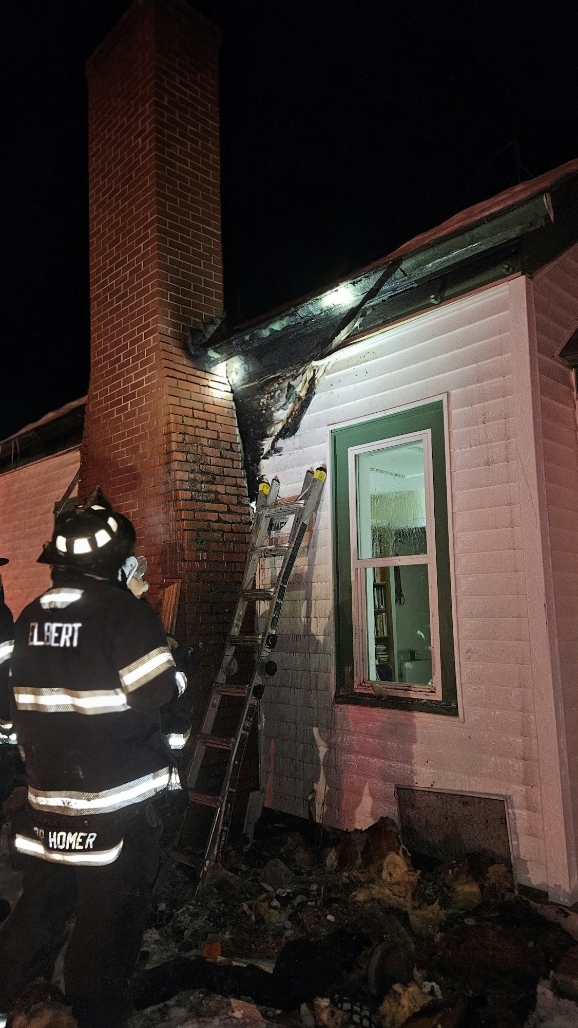 Firefighters inspecting fire damage on a building's exterior; ladder and burnt areas visible.