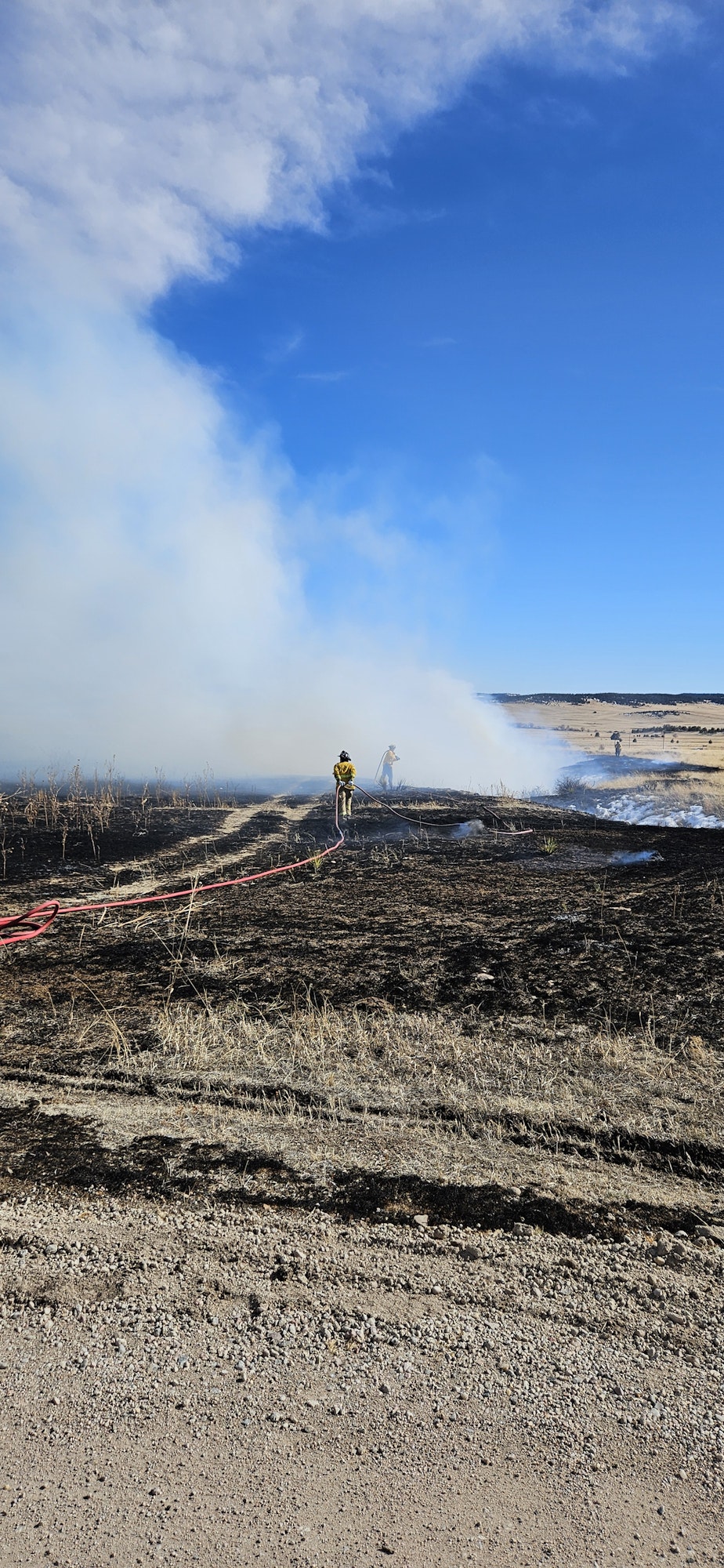 A person with a hose fights a grass fire, producing a large smoke cloud against a blue sky. (Image is rotated 90 degrees.)